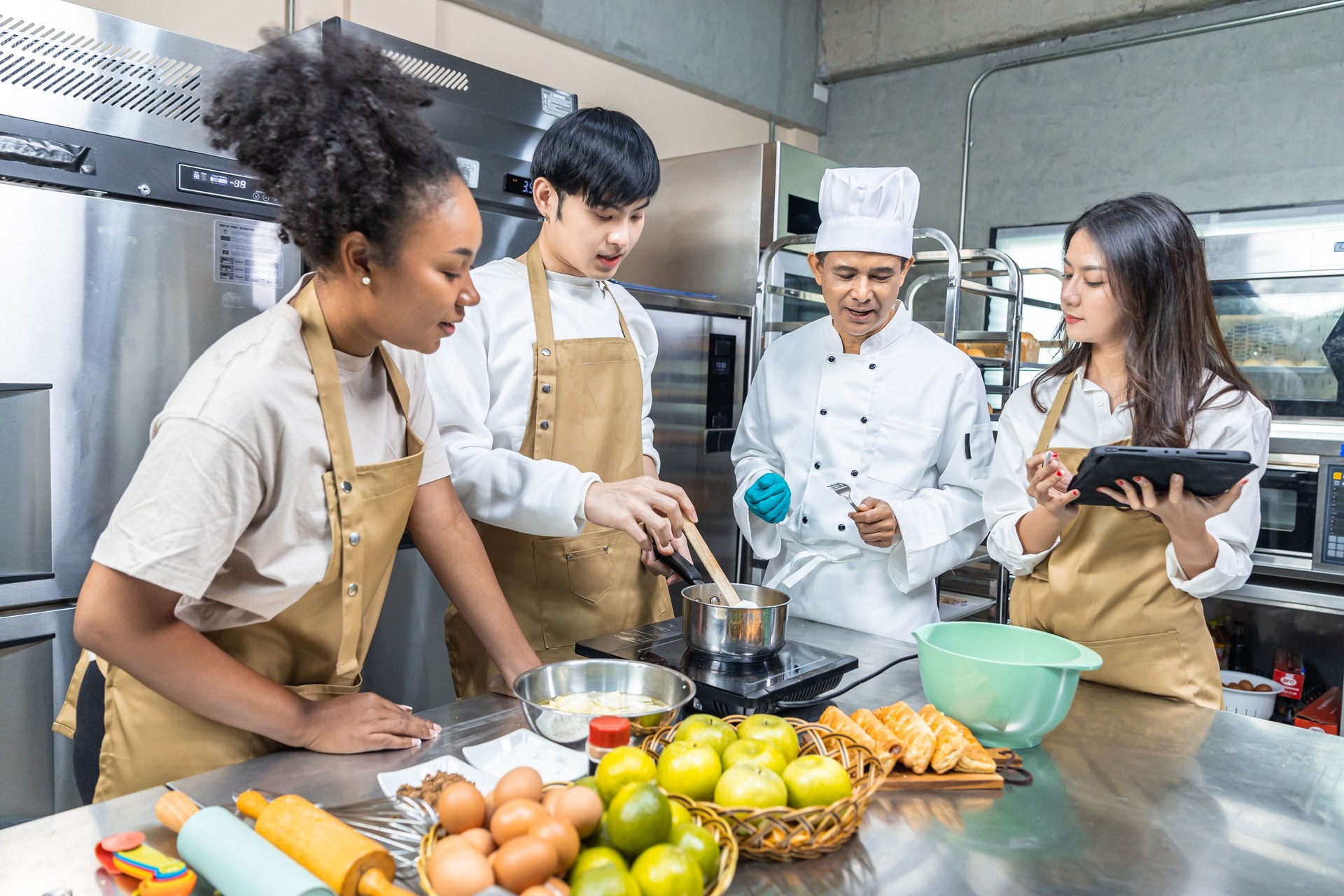 Students In Cookery Class Mixing Ingredients For Recipe In Kitchen.Male And Female young  Students With chef Teacher Preparing Ingredients For Dish In Kitchen Cookery Class.